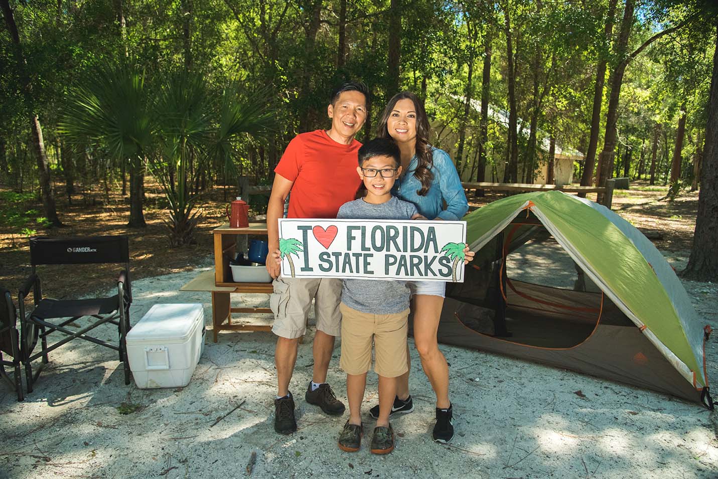A family of three holds up a sign reading “I love Florida State Parks” in front of a campsite. 
